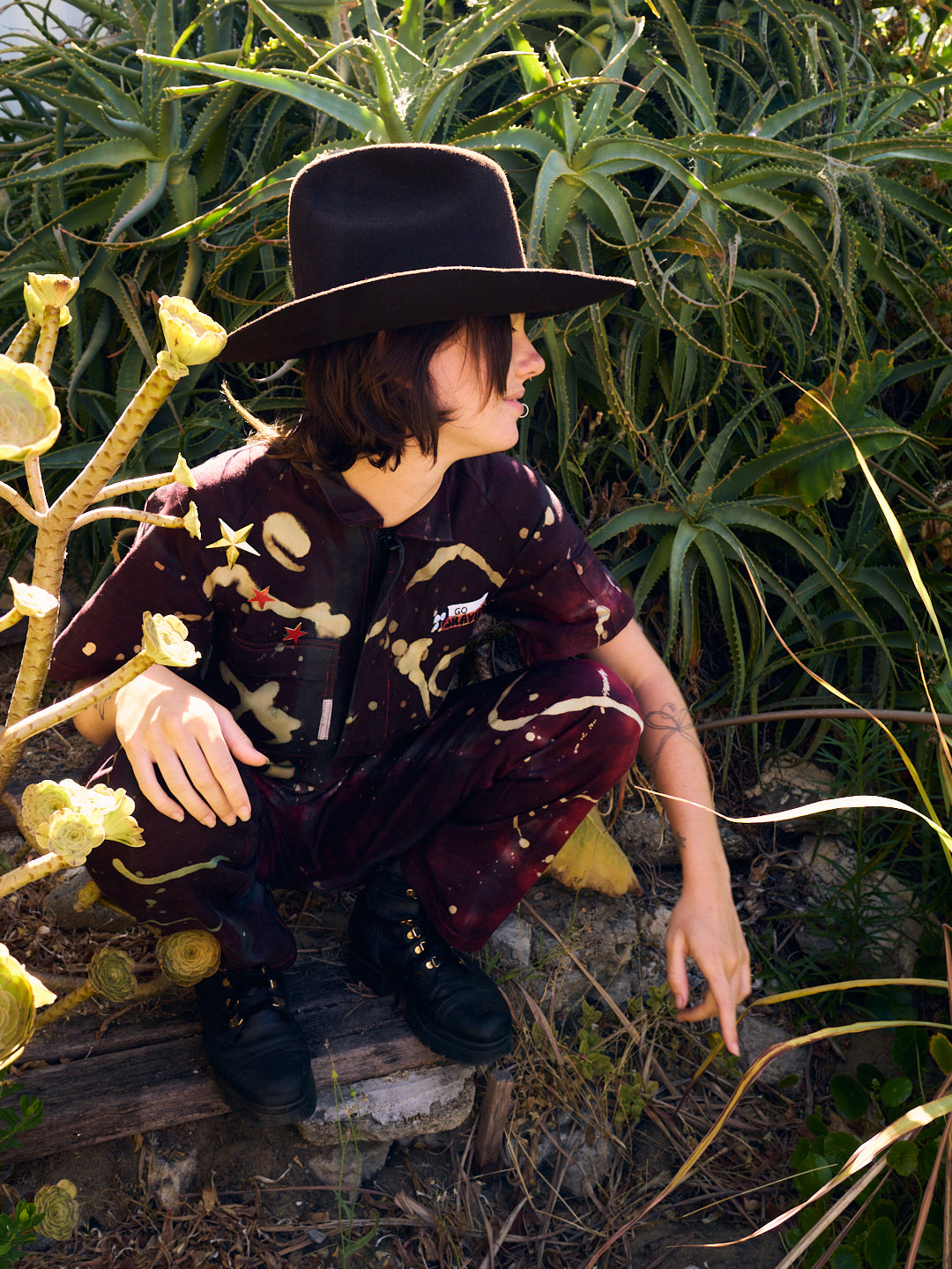 woman crouching on steps wearing cowboy hat and short sleeved overalls with galactic pattern stars and a badge with GO gravity on chest