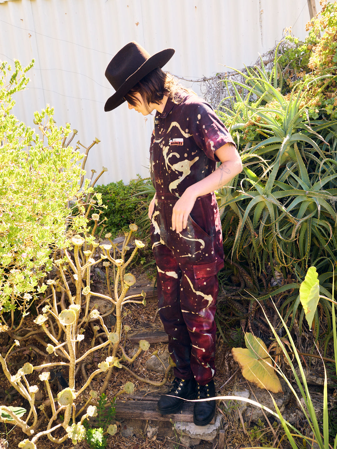 side view of woman wearing bleached and dyed maroon short sleeved jumpsuit and brown cowboy hat